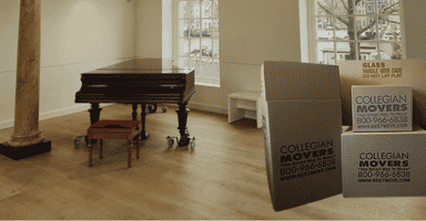 Baby grand piano on piano dolly in empty room with Collegian Movers boxes.